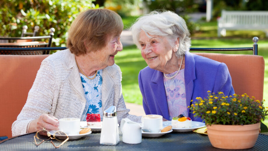 Two elderly women smiling and enjoying coffee together outdoors, symbolizing the importance of friendship and connection for longevity.